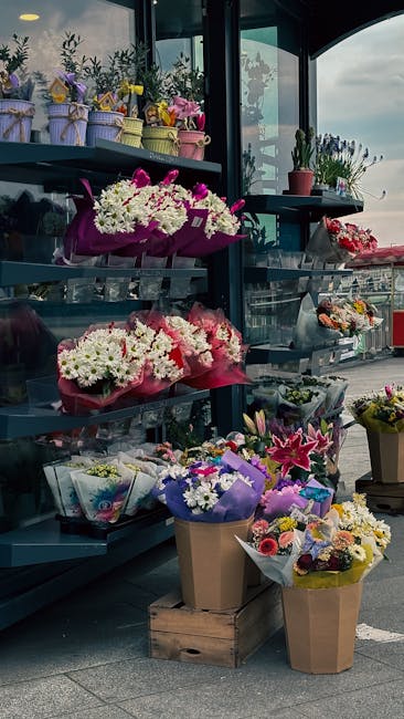 An outdoor floral display at a flower shop featuring a variety of fresh flower arrangements and bouquets. The arrangement on the lower shelf includes vibrant pink and white carnations wrapped in pink