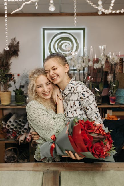 Two young women with light skin sharing a joyful moment in a floral shop, one holding a vibrant bouquet of red roses wrapped in grey and red paper with a satin ribbon. The woman on the left has curly