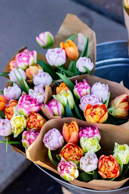 A vibrant bouquet of fresh tulips featuring shades of pink, orange, yellow, and white, arranged with green leaves and stems. The flowers are wrapped in light brown kraft paper and placed in a black co
