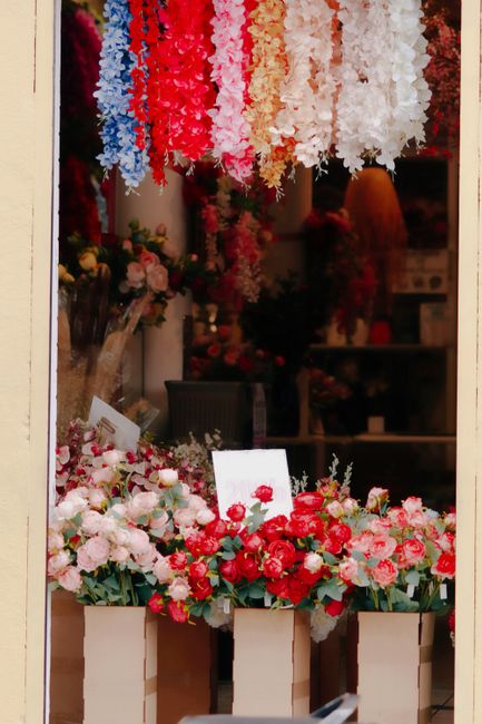 A flower shop display featuring vibrant floral arrangements and hanging floral garlands. In the foreground, there are three white boxes filled with fresh pink, red, and white roses, along with other m