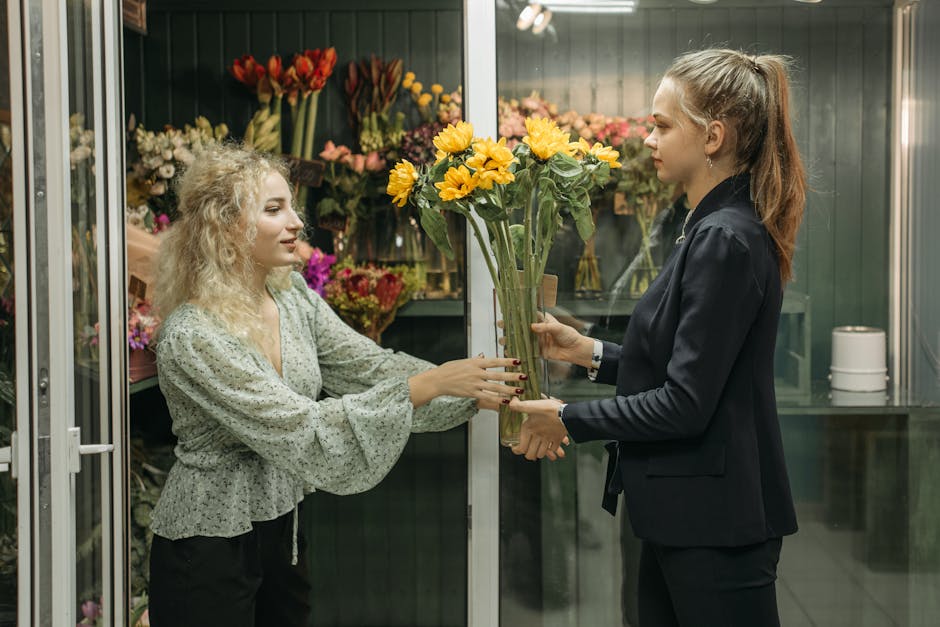 A woman with curly blonde hair and a light green floral blouse is handing over a bouquet of bright yellow sunflowers with green stems and leaves to a florist in a shop. The florist, with straight brow