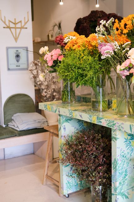 A display of vibrant fresh flowers arranged in clear glass vases on a floral-patterned table inside a flower shop. The bouquet includes large sunflowers with bright yellow petals, pink and white daisi