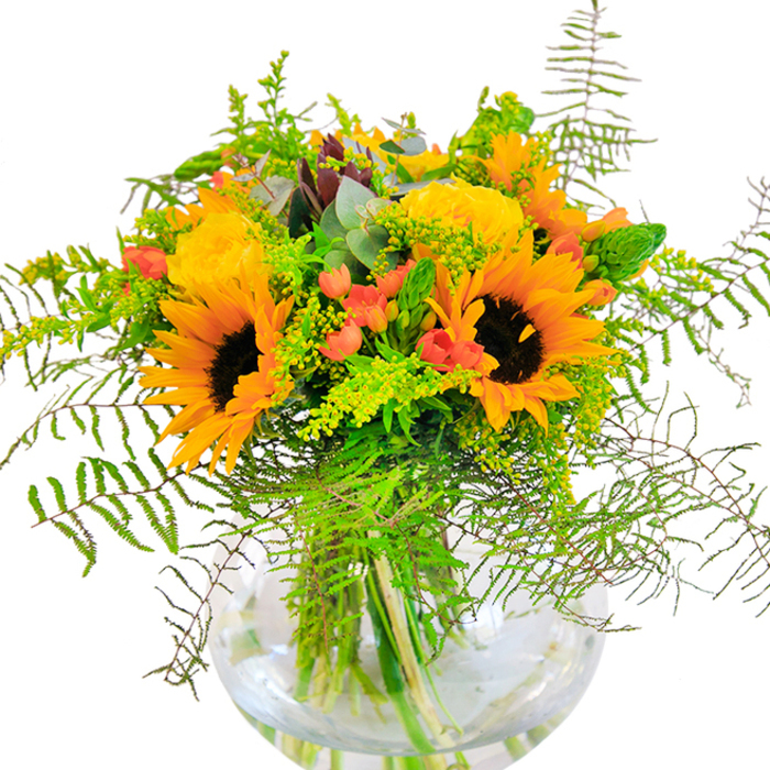 Instantly evoking the optimism of a sunlit afternoon in Hither Green, this artful bouquet pairs the brightness of yellow roses and sunflowers-a duo that radiates vitality-with playful touches of nature's wild elegance. The sunflowers, with their generous gold petals and deep cocoa centers, draw the eye, while tightly furled garden roses provide softness and tradition. Woven into their embrace are vibrant red Hypericum berries, their glossy surfaces gleaming against springy, deep green foliage reminiscent of the leafy serenity found around Manor Park. The bouquet's lush composition is layered intentionally: delicate ferns cascade downward, giving texture and movement, alongside airy spikes of solidago and slender green fillers. Eucalyptus leaves, in a subtle blue wash, peek through to add depth and a gentle fragrance. Contained within a round, clear glass vase, the arrangement's stems tangle elegantly in water-the transparency emphasizing freshness and craftsmanship. Light, hinting at a summer's close, plays across the blooms, casting gentle shadows that shift with time of day. Whether adorning a local Hither Green home, serving as a joyful centerpiece at a family gathering, or arriving as a thoughtful birthday or anniversary gift, this bouquet channels the friendly, vibrant character of SE13, connecting hearts and spaces with each petal.